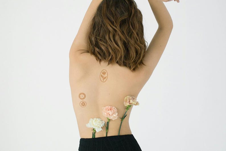Puede incluir: Un conjunto de tatuajes temporales marrones con un tulip&aacute;n en un marco ovalado y dos peque&ntilde;os dise&ntilde;os florales circulares sobre la piel, acompa&ntilde;ados de tres claveles frescos en blanco, rosa claro y melocot&oacute;n metidos en ropa negra.