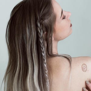 Puede incluir: Un tatuaje temporal que presenta un peque&ntilde;o dise&ntilde;o circular con una ilustraci&oacute;n floral blanca sobre un fondo marr&oacute;n claro, aplicado en la zona superior del hombro. El dise&ntilde;o muestra una flor sencilla con p&eacute;talos y tallo dentro de un borde circular.