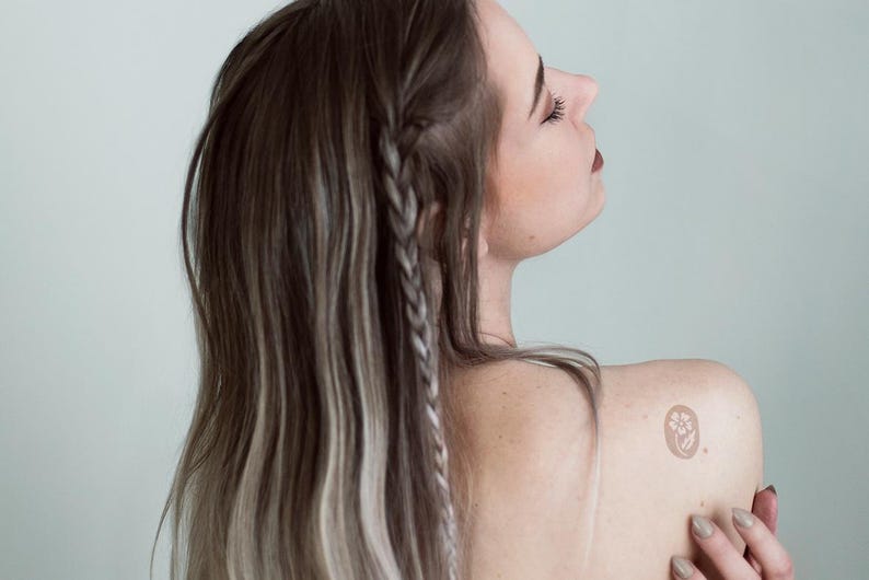 Puede incluir: Un tatuaje temporal que presenta un peque&ntilde;o dise&ntilde;o circular con una ilustraci&oacute;n floral blanca sobre un fondo marr&oacute;n claro, aplicado en la zona superior del hombro. El dise&ntilde;o muestra una flor sencilla con p&eacute;talos y tallo dentro de un borde circular.