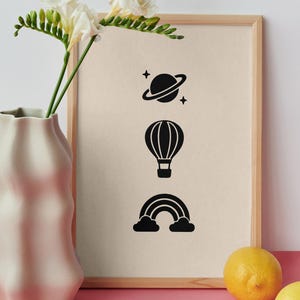 May include: A framed print featuring three black icons arranged vertically: a planet with rings and stars, a hot air balloon, and a rainbow with clouds. The print is displayed in a light wood frame next to a white vase with flowers and two yellow lemons.