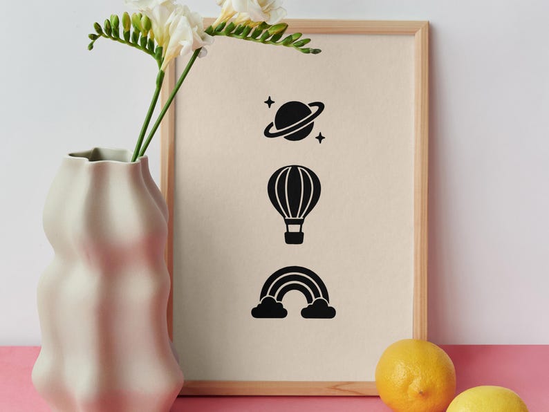 May include: A framed print featuring three black icons arranged vertically: a planet with rings and stars, a hot air balloon, and a rainbow with clouds. The print is displayed in a light wood frame next to a white vase with flowers and two yellow lemons.