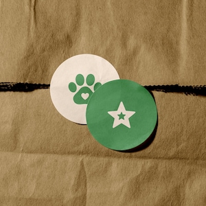 May include: Two circular stickers are layered on top of each other. The bottom sticker is white with a green paw print icon featuring a small heart in the center. The top sticker is green with a white five-pointed star icon in the center.