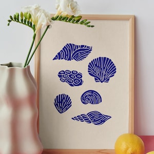 Puede incluir: Una l&aacute;mina enmarcada que presenta una colecci&oacute;n de seis ilustraciones en azul oscuro de varios tipos de conchas marinas y formaciones de coral dispuestas sobre un fondo beige claro. La obra est&aacute; en un marco de madera clara.