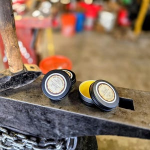 May include: Several small, round, black tins of product with white and brown labels are displayed on a metal surface. One tin is open, revealing a yellow substance. A hammer rests on the surface, and a chain is visible in the foreground.