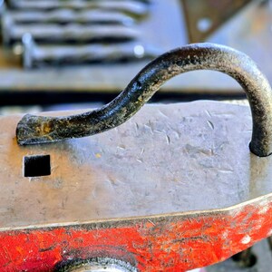 Peut inclure: Gros plan d'une enclume de forgeron avec une poign&eacute;e m&eacute;tallique incurv&eacute;e. L'enclume est principalement grise avec une base rouge. Un trou carr&eacute; est visible sur la surface de l'enclume. La poign&eacute;e est sombre et textur&eacute;e.