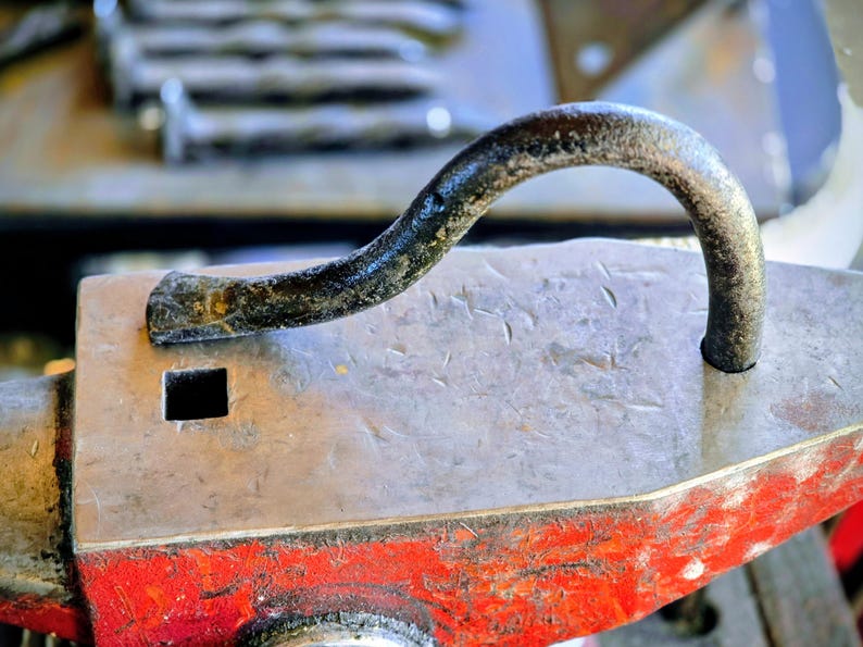 Peut inclure: Gros plan d'une enclume de forgeron avec une poign&eacute;e m&eacute;tallique incurv&eacute;e. L'enclume est principalement grise avec une base rouge. Un trou carr&eacute; est visible sur la surface de l'enclume. La poign&eacute;e est sombre et textur&eacute;e.