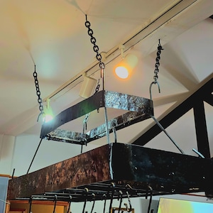 May include: A wrought iron pot rack suspended from the ceiling by chains. The rectangular rack has a lower shelf with hooks for hanging pots and pans. Two spotlights are mounted on a track above the rack.