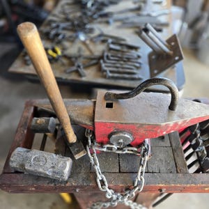 Peut inclure: Un &eacute;tabli de forgeron avec une enclume rouge et argent&eacute;e, un marteau avec un manche en bois et une masse. Une cha&icirc;ne fixe l'enclume &agrave; une base en bois. Divers outils sont visibles en arri&egrave;re-plan, sugg&eacute;rant un atelier.