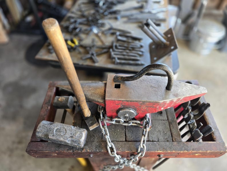 Peut inclure: Un &eacute;tabli de forgeron avec une enclume rouge et argent&eacute;e, un marteau avec un manche en bois et une masse. Une cha&icirc;ne fixe l'enclume &agrave; une base en bois. Divers outils sont visibles en arri&egrave;re-plan, sugg&eacute;rant un atelier.