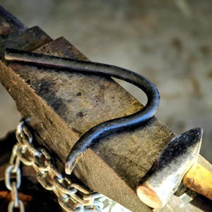 Peut inclure: Enclume de forgeron avec une pi&egrave;ce m&eacute;tallique incurv&eacute;e pos&eacute;e dessus. Un marteau avec un manche en bois et une cha&icirc;ne sont &eacute;galement visibles. L'enclume est en m&eacute;tal sombre et vieilli, sugg&eacute;rant un atelier.