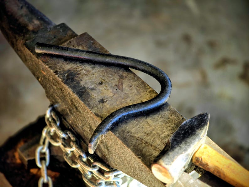 Peut inclure: Enclume de forgeron avec une pi&egrave;ce m&eacute;tallique incurv&eacute;e pos&eacute;e dessus. Un marteau avec un manche en bois et une cha&icirc;ne sont &eacute;galement visibles. L'enclume est en m&eacute;tal sombre et vieilli, sugg&eacute;rant un atelier.