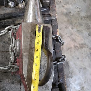 Peut inclure: Une enclume en m&eacute;tal avec une corne conique et une base rectangulaire, avec un c&ocirc;t&eacute; peint en rouge. Un m&egrave;tre ruban jaune indique que l'enclume mesure environ 25 cm de haut. Une cha&icirc;ne est attach&eacute;e &agrave; l'enclume.