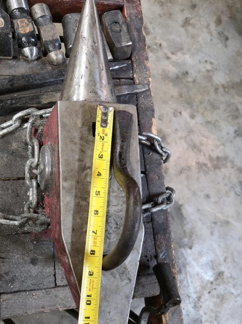 Peut inclure: Une enclume en m&eacute;tal avec une corne conique et une base rectangulaire, avec un c&ocirc;t&eacute; peint en rouge. Un m&egrave;tre ruban jaune indique que l'enclume mesure environ 25 cm de haut. Une cha&icirc;ne est attach&eacute;e &agrave; l'enclume.