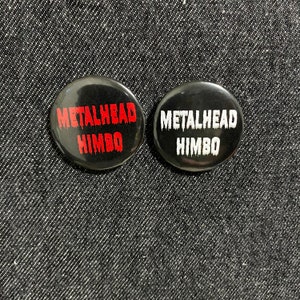 May include: Two black button badges with the text "Metalhead Himbo" in red and white lettering.