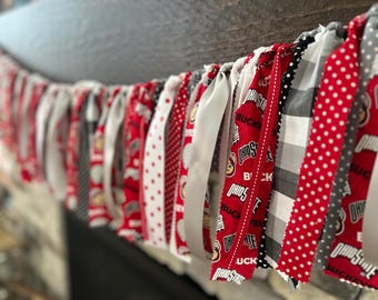 Puede incluir: Guirnalda de tela festiva con cintas estampadas rojas, blancas y grises alternas. Los patrones incluyen lunares, rayas y logotipos de Ohio State Buckeyes. Ideal para fiestas temáticas o días de partido.
