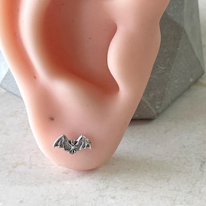 May include: A silver bat stud earring on a model ear. The earring is small and delicate, with a detailed bat design.