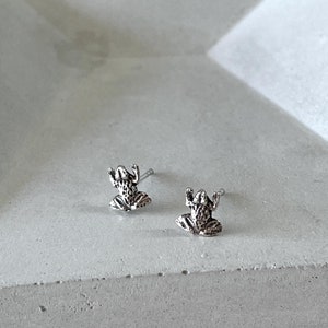 May include: A pair of silver frog stud earrings on a grey concrete tray.