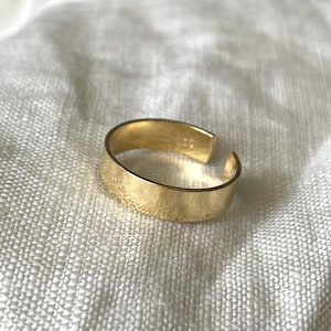 Peut inclure: Une bague ajustable dorée à la surface lisse et plate. La bague est ouverte d'un côté et a une forme légèrement incurvée.