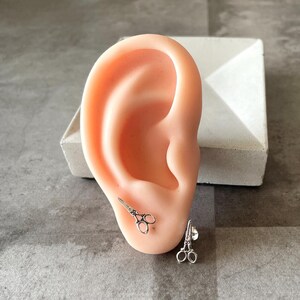 May include: A pair of silver earrings shaped like scissors. The earrings are shown on a model's ear.