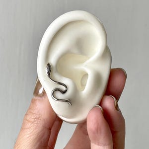 May include: A silver snake ear cuff is shown on a white ear model. The ear cuff wraps around the ear, creating a unique accessory. The snake design adds an edgy elegance to the jewelry. The ear cuff is a stylish piece.