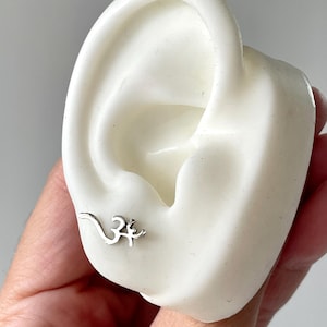 May include: A silver-colored stud earring in the shape of the Om symbol is displayed on a white ear model. The earring is small and detailed, with a curved design. The ear model is held by a hand with a light skin tone.