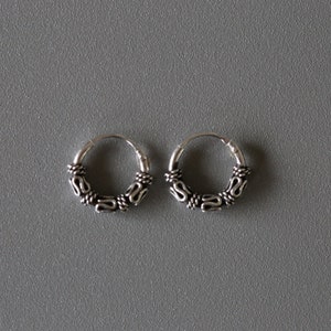 Puede incluir: Un par de pendientes de aro de plata con un diseño detallado e intrincado. Los aros son pequeños y delicados.