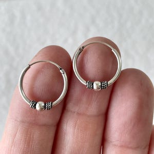 May include: A pair of silver hoop earrings, each featuring a smooth central bead flanked by two small, textured, rope-patterned silver accents. The hoops are circular with a thin wire construction and a simple hinge closure mechanism.