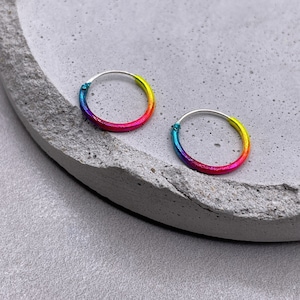 Puede incluir: Un par de pendientes de aro de colores arcoíris. Los pendientes son de plata y tienen un degradado de colores que va del azul al amarillo.