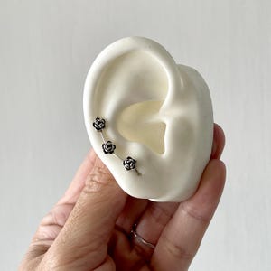 May include: A silver ear cuff featuring three small rose designs. The ear cuff is shown on a white ear model, held by a hand. The roses are detailed and add a touch of floral elegance to the jewelry.
