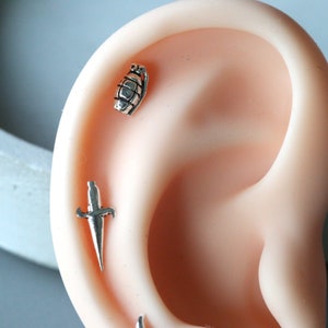 May include: A close-up of a person's ear with two silver earrings. The earring on the top is shaped like a grenade, and the earring on the bottom is shaped like a dagger.
