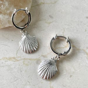 May include: A pair of silver hoop earrings with seashell charms. The earrings have a polished finish and a detailed seashell design, perfect for a beach-themed look. The earrings are shown on a light-colored surface.