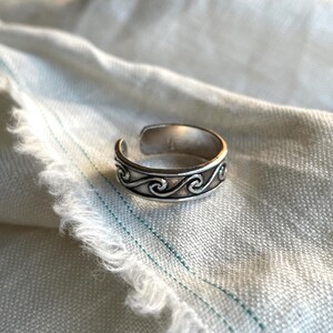 Può includere: Anello per dita dei piedi in argento con un design a onde. L'anello è regolabile e ha una finitura liscia.