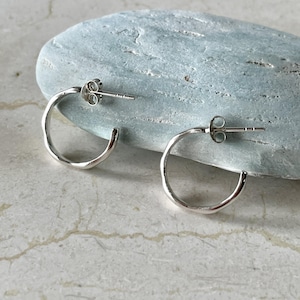 Puede incluir: Un par de pendientes de aro pequeños y plateados. Los pendientes tienen un aspecto texturizado y martillado y un cierre de poste y mariposa. Se exhiben sobre una superficie de piedra azul claro, mostrando su diseño simple y elegante. Un accesorio de joyería clásico.