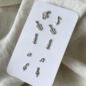 May include: A collection of silver-colored musical-themed earrings on a white card. The earrings include treble clefs, eighth notes, and miniature guitars and violins. The earrings are small and detailed, perfect for music lovers.