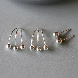 Puede incluir: Un conjunto de tres pendientes de plata. Dos pendientes tienen un diseño curvo con tres bolas de plata en el extremo. El otro pendiente es un simple pendiente de botón con una sola bola de plata.