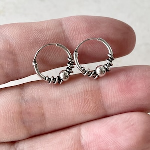 May include: A pair of small silver hoop earrings, each featuring a polished spherical bead at the center flanked by coiled wire detailing. The hoops are constructed from thin, rounded silver wire, showcasing a metallic finish with dark oxidation in the crevices of the coiled sections, providing a clear view of the intricate metalwork design.