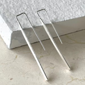 May include: A pair of silver bar earrings. Each earring features a long, rectangular bar that curves at the top to form a hook. The earrings are displayed on a textured, off-white surface.