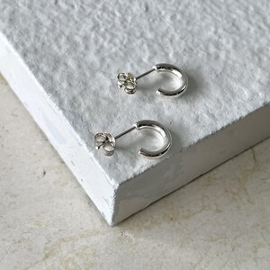 May include: A pair of small, shiny silver hoop earrings. Each earring has a curved, semi-circular design with a post and butterfly backing. The earrings are displayed on a textured, white surface.