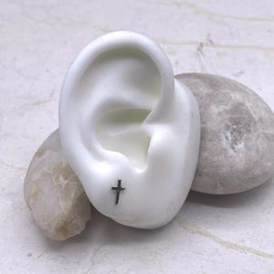 May include: A small, silver-colored cross stud earring is displayed on a white ear model. The earring is a simple, minimalist design. The ear model is positioned on a light-colored, textured surface with two smooth, gray stones.