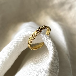 Könnte beinhalten: Ein goldener geflochtener Ring mit einem schlichten Design. Der Ring ist aus dickem, geflochtenem Draht gefertigt und hat eine glatte, polierte Oberfläche.