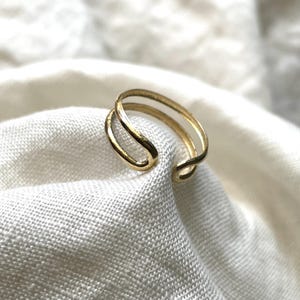 Könnte beinhalten: Ein goldfarbener Doppelring mit schlichtem, minimalistischem Design. Der Ring besteht aus zwei dünnen Bändern, die oben und unten miteinander verbunden sind und so einen einzigartigen und stilvollen Look erzeugen.