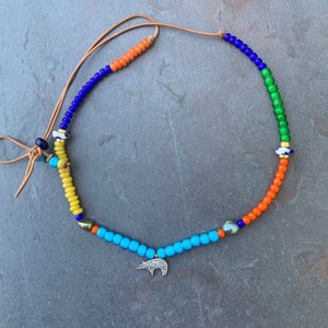 Puede incluir: Un collar de cuentas de colores con un dije de oso. El collar está hecho con cuentas azules, verdes, naranjas y amarillas. El dije de oso es de plata con un diseño de diamante.