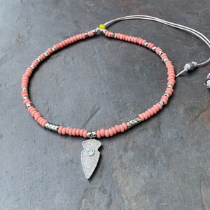 Puede incluir: Un collar de cuentas rosadas y plateadas con un colgante de punta de flecha plateada con un cristal transparente. El collar tiene un cordón blanco con una pequeña cuenta azul y blanca.