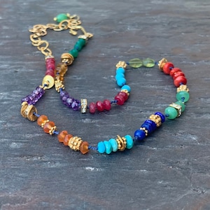 Puede incluir: Un collar de piedras preciosas arcoíris con detalles dorados y un cordón azul. El collar presenta cuentas de amatista, cornalina, rubí rojo, turquesa, lapislázuli y jade verde.