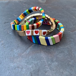 May include: Three colorful bracelets with gold accents and red heart charms. The bracelets are made of small, square tiles in various colors, including yellow, blue, green, red, and white.