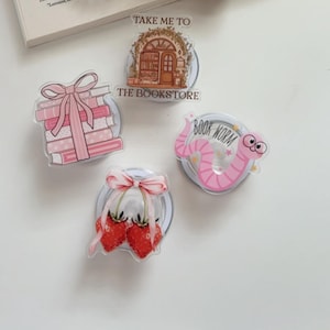 May include: A collection of decorative phone grips featuring various designs. Includes a pink bow, a pearl beaded wrist strap, and several pop sockets with images of a bookstore, books, strawberries, and a pink worm.
