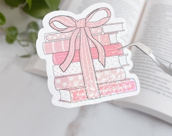 May include: A stack of pink books tied with a pink ribbon sticker. The books have various patterns, including floral and hearts. The sticker is held by tweezers in front of an open book and a plant.