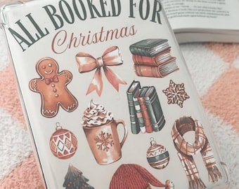May include: A white and clear phone case with the text "All Booked for Christmas" in green and brown lettering. The case is decorated with illustrations of a gingerbread man, a bow, a cup of hot chocolate, a Christmas tree, a hat, a scarf, and stacks of books.