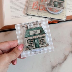 May include: A clear acrylic book shop-themed phone grip with a green roof and the text "BOOK SHOP." The grip is on a checkered background. The image also shows a library card-themed phone grip with the text "Ask me about Books."