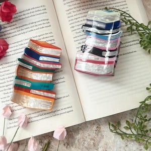 Op de afbeelding: Gestapelde haarclips in de vorm van boeken in verschillende kleuren, waaronder oranje, blauw en turquoise, met witte en parelmoer accenten. De clips liggen op een open boek met tekst en zijn omringd door roze bloemen.